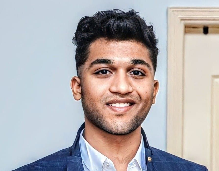 Person wearing a blue suit jacket and white shirt standing indoors near a door.