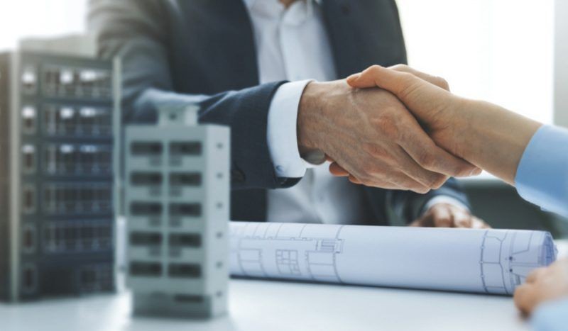 Two people shaking hands over a table with architectural blueprints and building models.